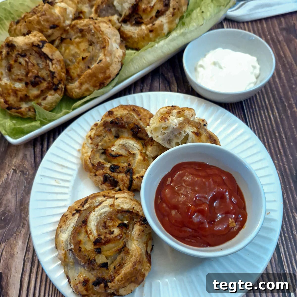 Savory Sausage and Caramelized Onion Pinwheel Appetizers 3 Sausage and onion pinwheels on a white plate with a small dish of tomato ketchup for dipping. There is a plate of sausage pinwheels in the background.