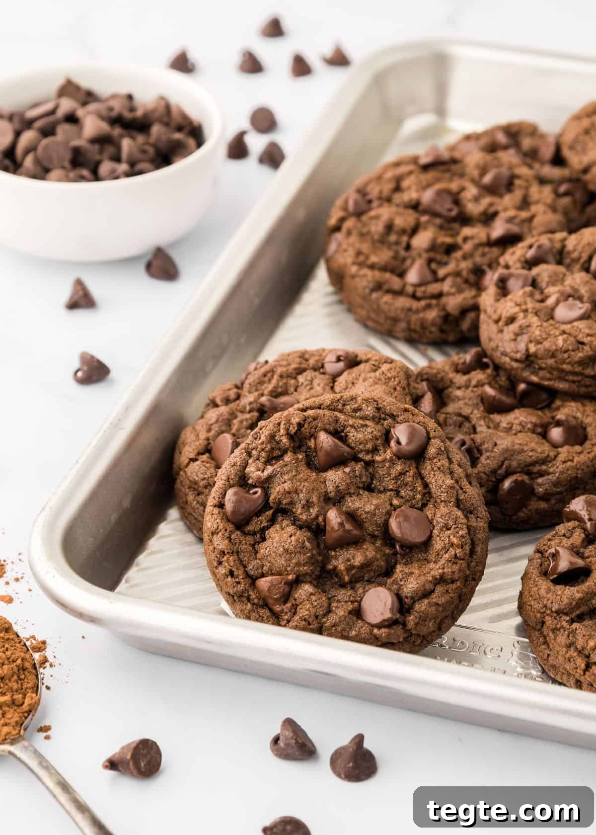 Ultimate Double Chocolate Cookies Recipe: Moist, Fudgy Bites 12 Chocolate chip cookies on a baking sheet on a counter with a spoonful of cocoa powder.