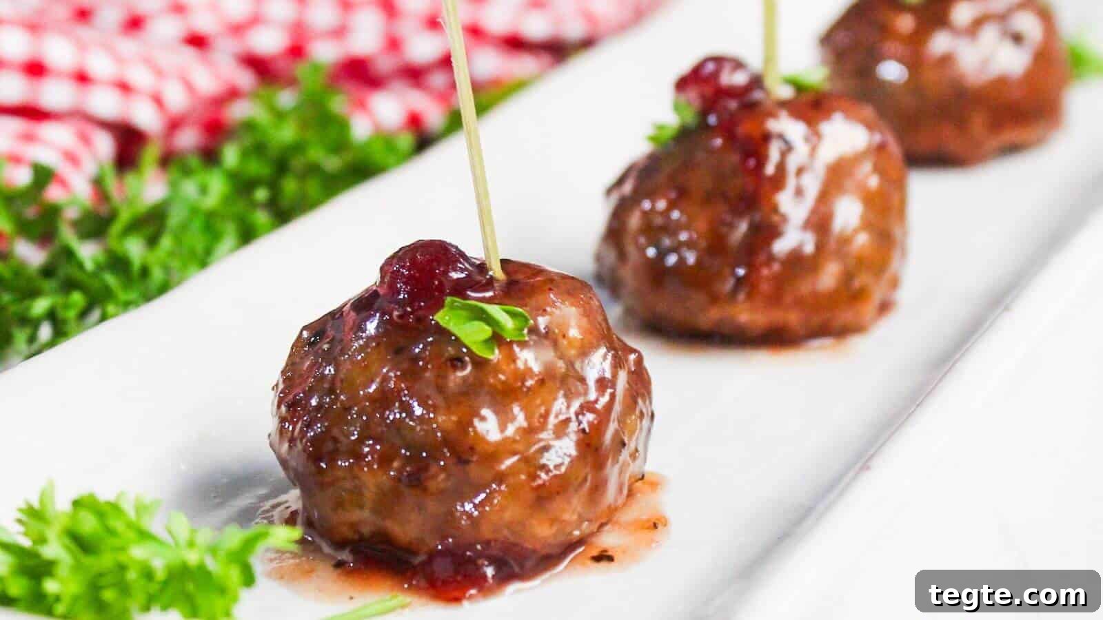 15 Creative Post-Thanksgiving Dinners to Transform Leftovers 4 Three glazed meatballs are arranged in a row on a white plate, each garnished with a stick and a small sprig of parsley. A red checkered cloth and some fresh parsley are in the background.
