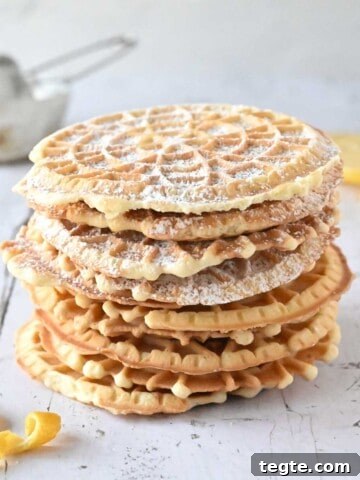 Lemon pizzelles stacked on top of each other on white wood with lemon in the background.