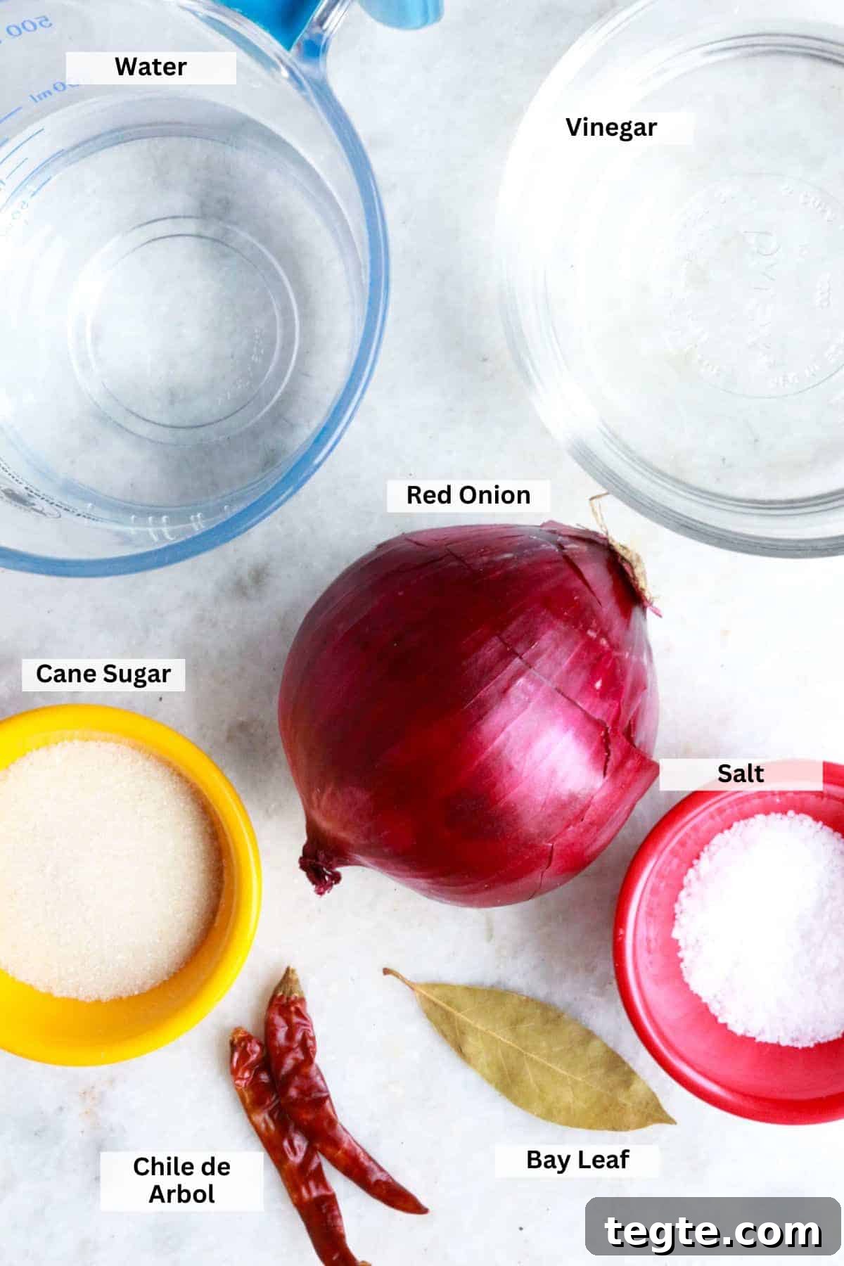Bowl of white vinegar, measuring cup with water, sugar, chiles de arbol, bay leaf, red onion and salt