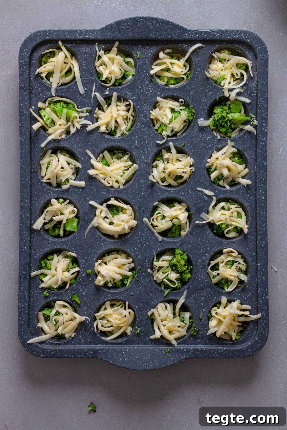 A mini muffin pan with shredded cheese and chopped broccoli.