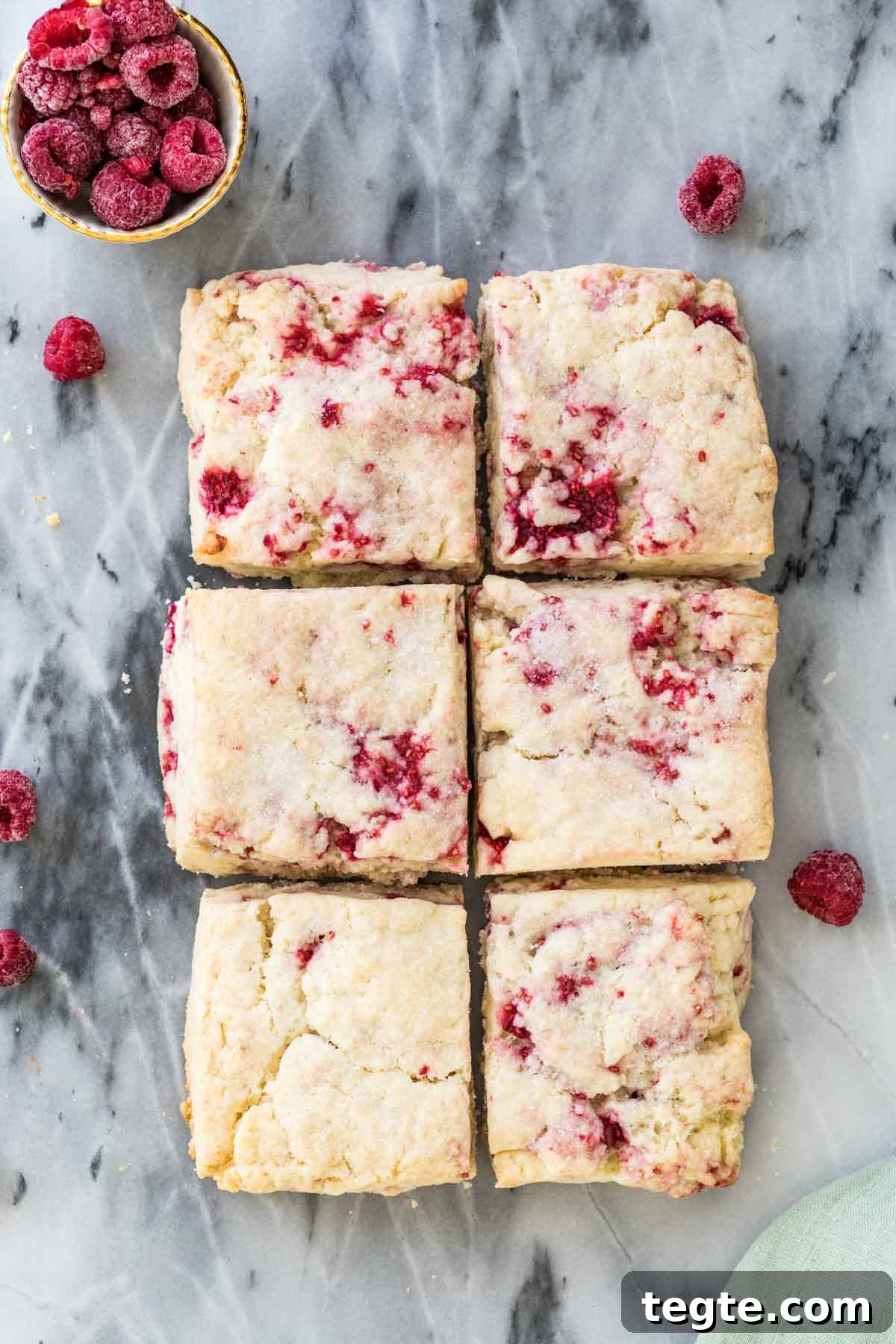Overhead shot of raspberry scones on gray marble next to a bowl of raspberries in the top left corner