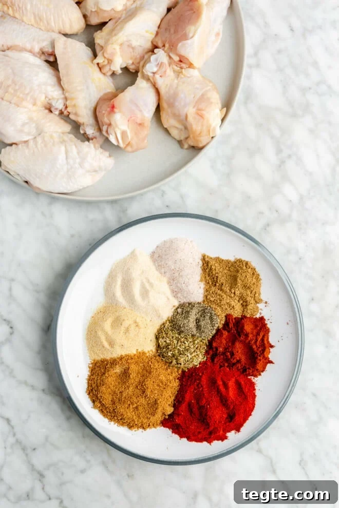 Crispy Dry Rub Chicken Wings Recipe for Perfect Flavor 4 There are two round plates. One is a white plate with a blue rim that has a mixture of individually scooped spices on the plate. The second is a grey plate that has raw chicken wings on the plate. Both are sitting on a grey and white marble surface.