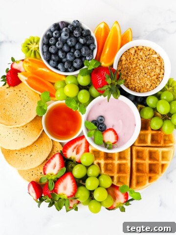 A round board filled with pancakes, waffles, fruit, yogurt, and syrup