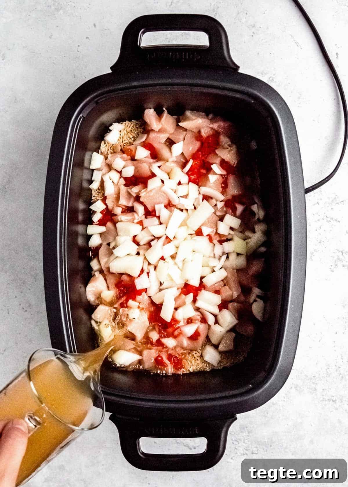 chicken broth being poured into black crockpot filled with cubed chicken, chopped onions, pimentos, and brown rice