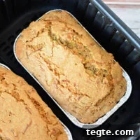 Two loaves of golden-brown zucchini bread in metal trays rest perfectly inside a sleek black air fryer basket.