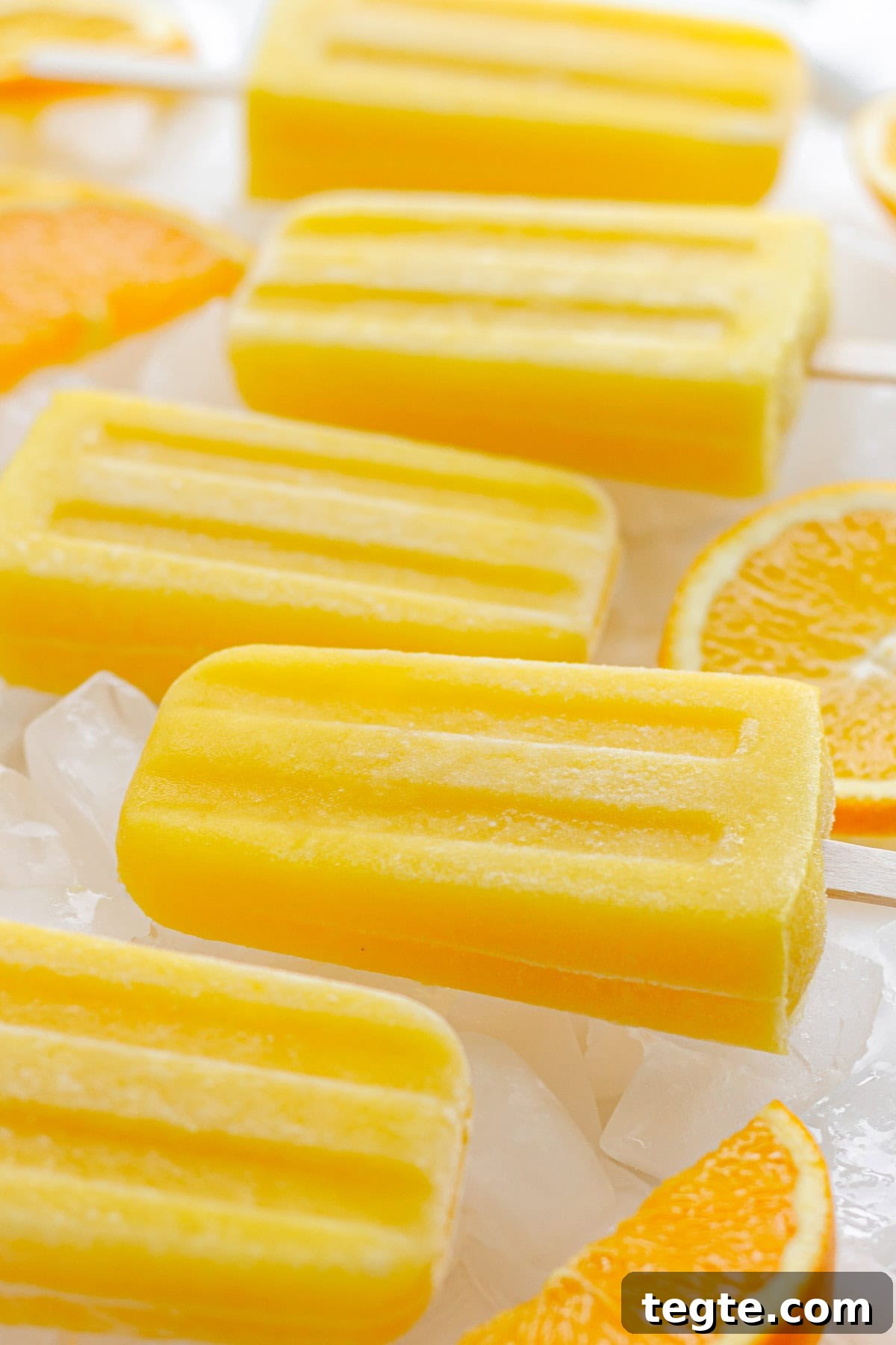 Refreshing Homemade Orange Popsicles Recipe 3 Orange popsicles lined up on ice cubes with some orange slices.