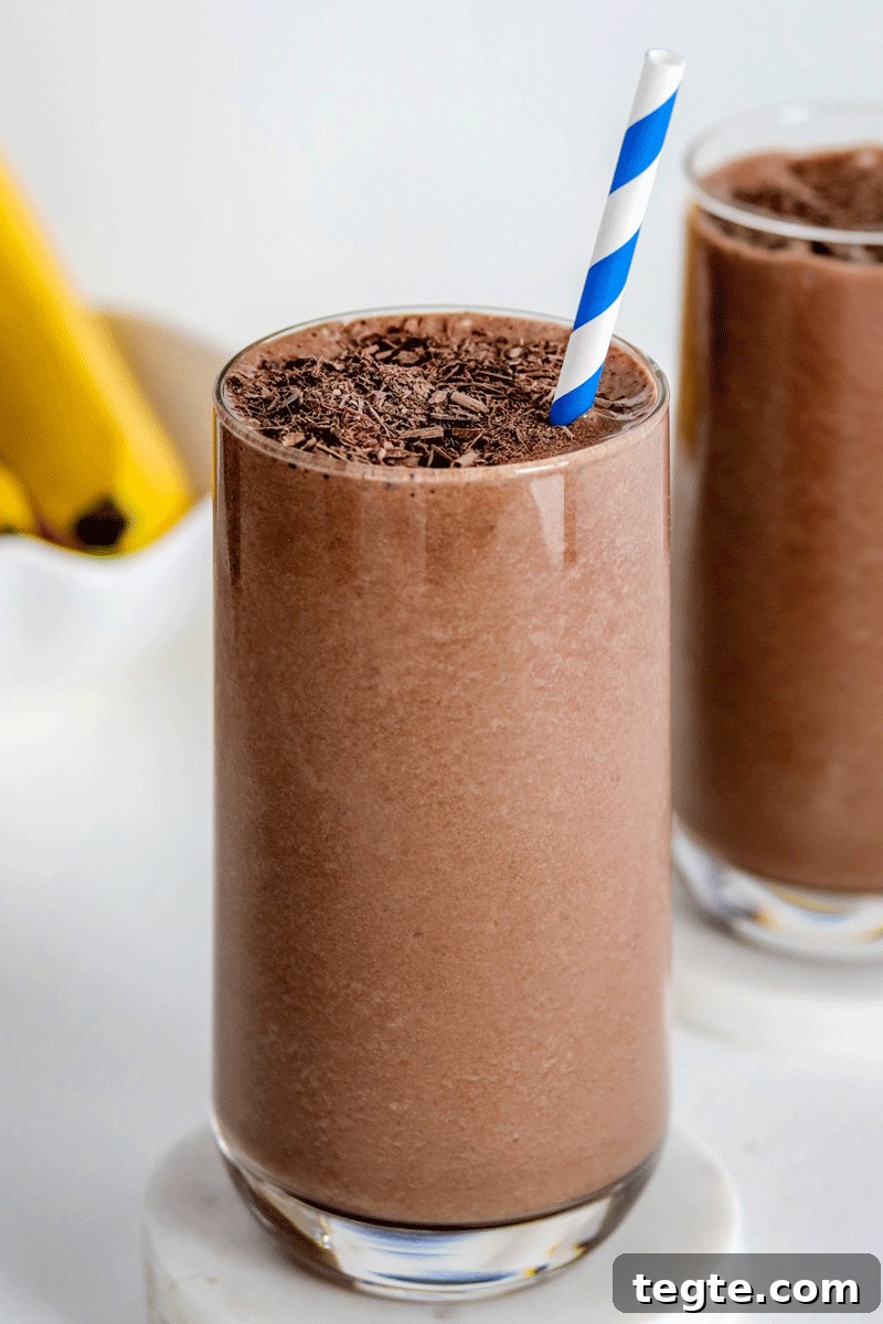 Two chocolate smoothies with a shaved chocolate on top and a bunch of bananas in the background