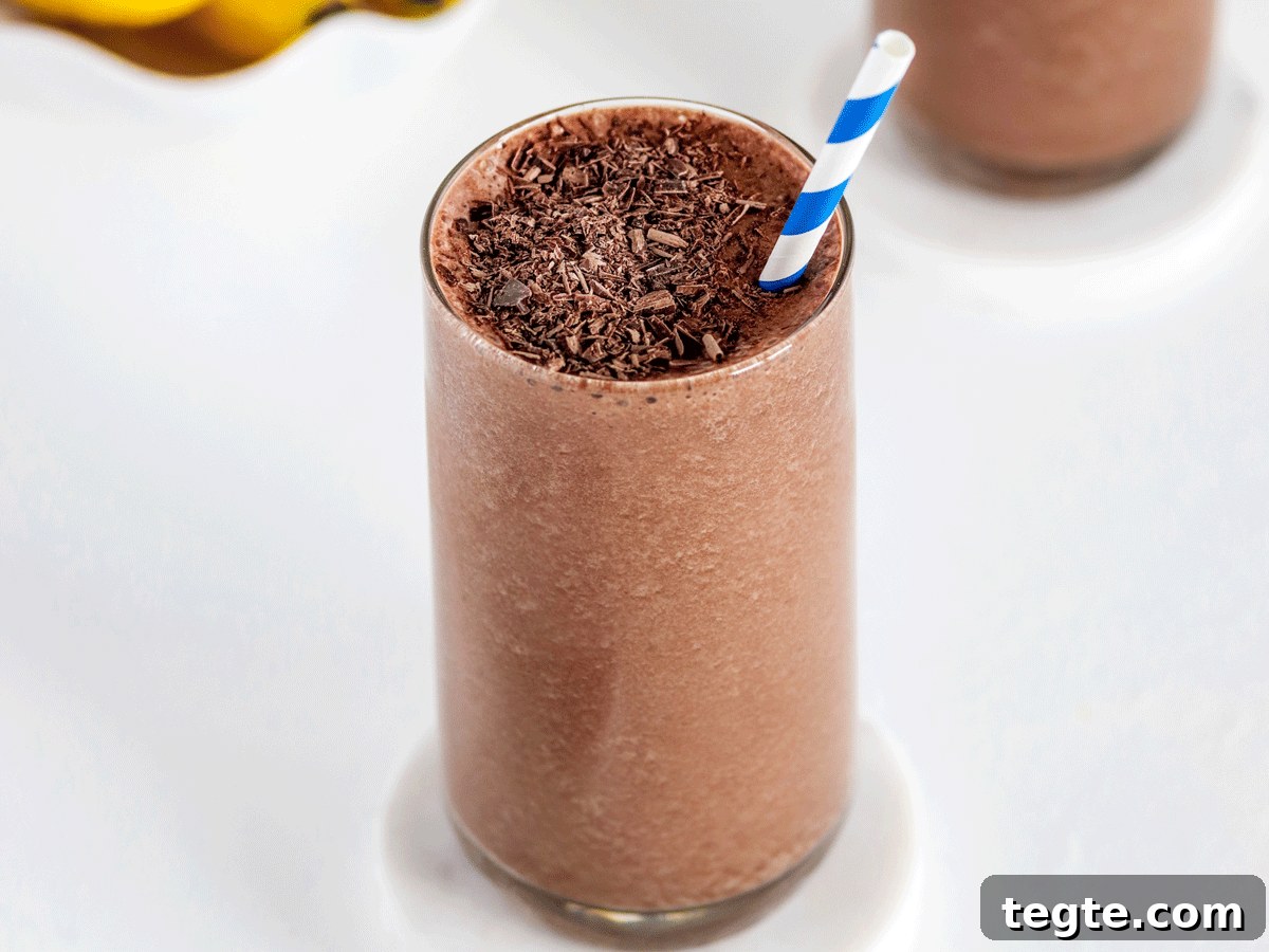 Two chocolate smoothies with a shaved chocolate on top and a bunch of bananas in the background