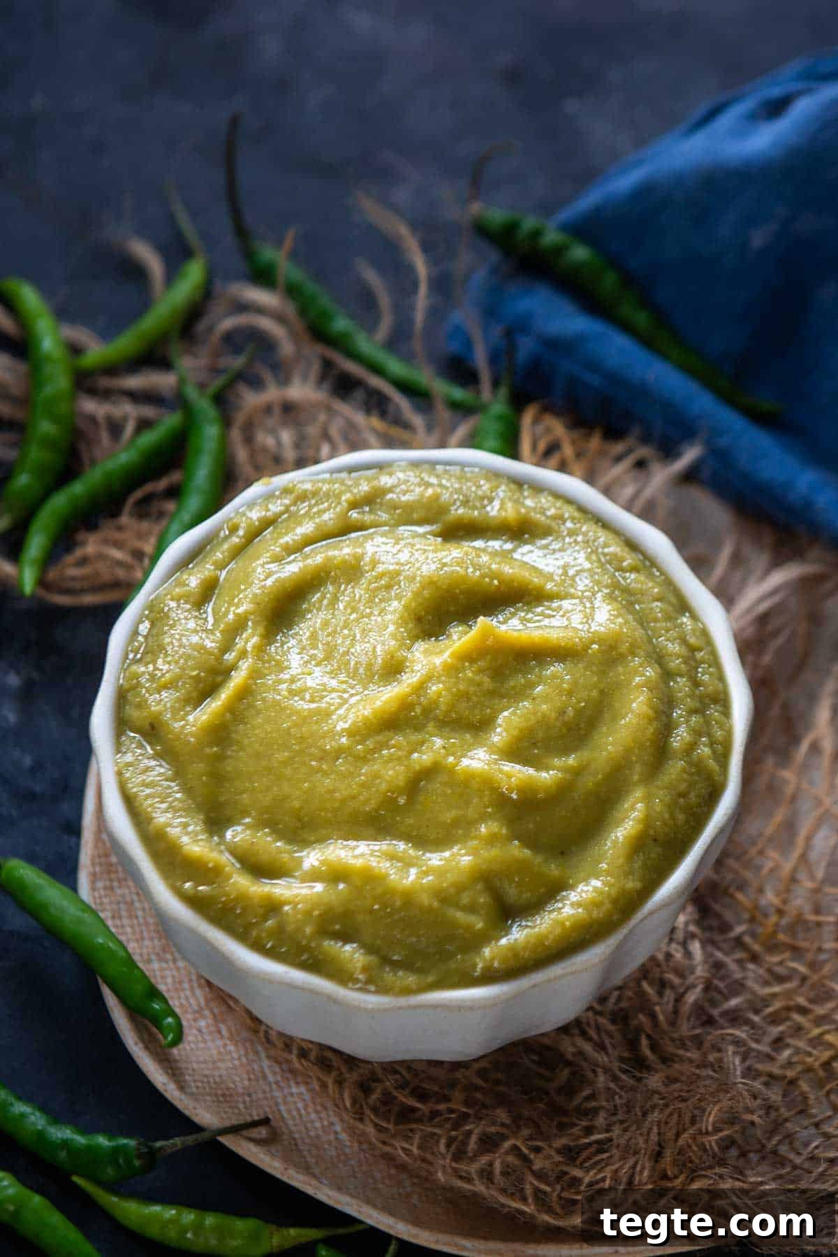 Indo-Chinese Green Chili Sauce Recipe for Spicy Dips and Stir-Fries 2 A white bowl filled with smooth Green Chilli Sauce sits on a piece of burlap, surrounded by whole green chilies on a dark surface, with a blue cloth in the background.