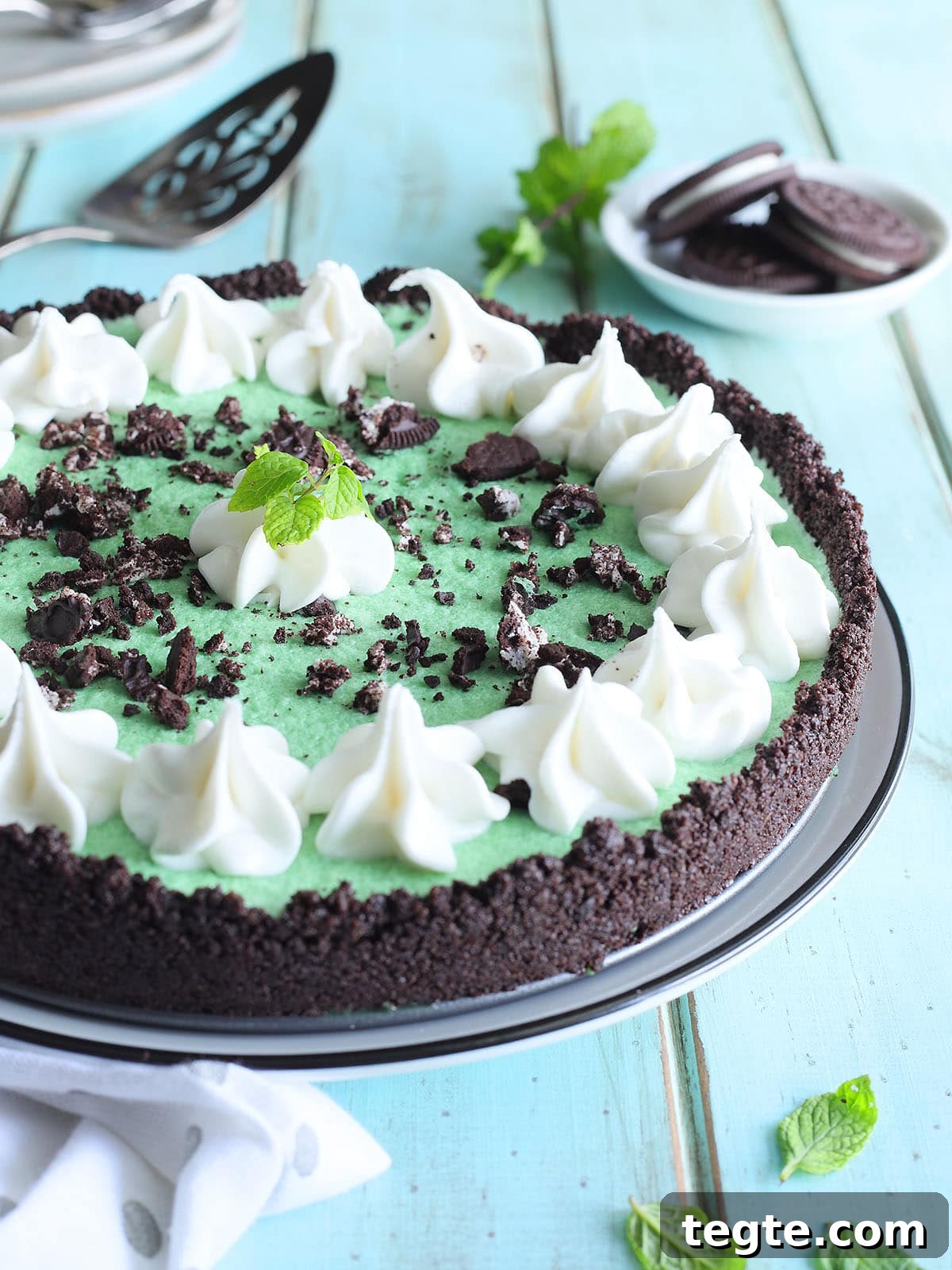 Classic Grasshopper Pie Recipe: Mint Chocolate Cream Pie 12 Grasshopper pie garnished with piped whipped cream, crushed cookies, and a mint sprig. A small dish of chocolate sandwich cookies and additional fresh mint are in the background.