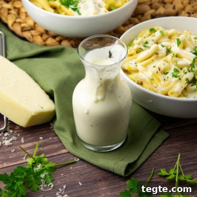 Creamy Asiago Sauce for Pasta and Vegetables 7 a glass carafe of white cheese sauce in front of a bowl of pasta