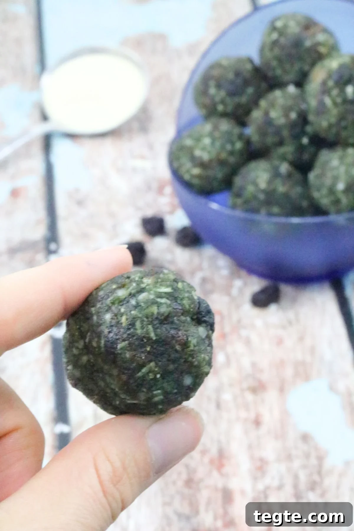 Energizing Spirulina Protein Balls (Gluten-Free) 2 Two fingers holding up a superfood energy ball with more balls in a blue bowl behind them.