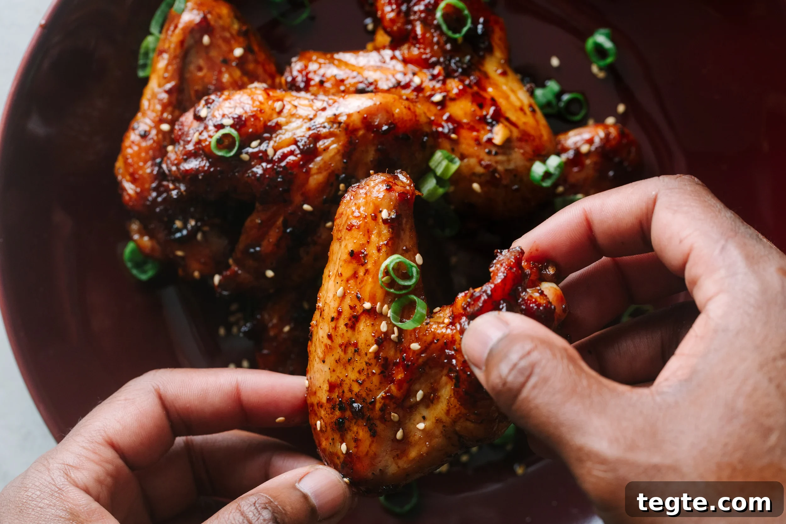 Asian Chili Garlic Chicken Wings Recipe 16 Hands holding Asian chicken wings garnished with sesame seeds and green onions.